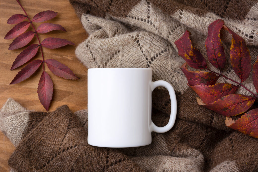 Maqueta de una taza de café blanca con una manta de punto y hojas rojas de serbal otoñales.